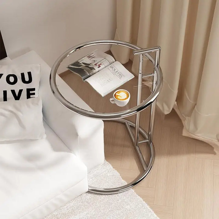 Small round glass table with a metal frame next to a bed with pillows and a blanket.